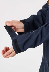 Roke, ki nastavljajo črni ježek na manšeti temno modre jakne z dolgimi rokavi na enostavni beli podlagi.