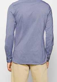 Camicia a maniche lunghe in tessuto blu scuro con una texture liscia, caratterizzata da polsini con bottoni e colletto classico, indossata con pantaloni beige chiaro.