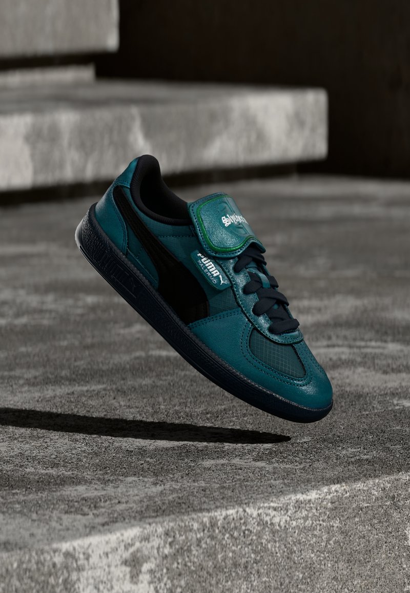 Baskets Puma bleu sarcelle et noir avec lacets et bande Velcro flottant au-dessus de marches en béton dans un cadre urbain.
