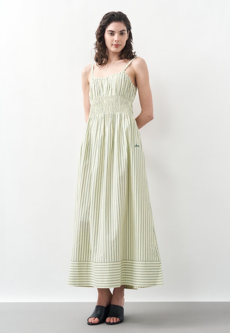 Femme portant une robe maxi sans manches à rayures vert clair et blanc avec un corsage smocké et des chaussures noires ouvertes à enfiler, debout contre un mur blanc.