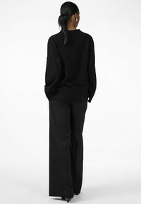 Maglione nero oversize con maniche lunghe, abbinato a pantaloni neri a gamba larga. Entrambi i capi presentano una consistenza liscia e un design minimale.