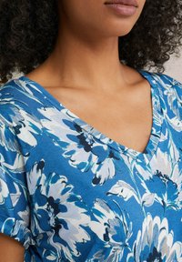 Zils ziedu tops ar V veida kakla izgriezumu, īsām piedurknēm un vieglu audumu. Dizainā ir lieli balti un tumši zili ziedi.