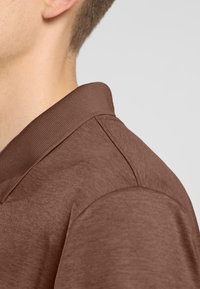 Polo marron avec un col côtelé et une texture lisse, mettant en avant un design ajusté avec une couture d'épaule visible et des détails en tissu.