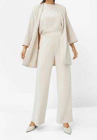Béžový třídílný outfit skládající se z volné, otevřené bundy s širokými rukávy, přiléhavého topu a kalhot s širokými nohavicemi. Hladká, jemná textura.