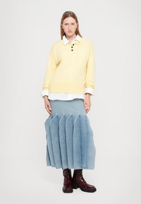 Maglione giallo a maglia con tre bottoni, indossato sopra una camicia bianca. Gonna a pieghe in denim blu con orlo grezzo. Stivali bicolore lucidi color bordeaux.