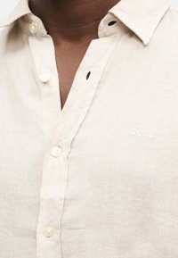 Gros plan sur une chemise en lin beige clair avec des boutons, un col et une broderie discrète "BOSS" sur la poitrine, portée par une personne à la peau foncée.