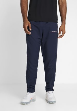 Mężczyzna w granatowych spodniach Tommy Sport i białych butach sportowych, stojący na tle jednolitego białego tła.