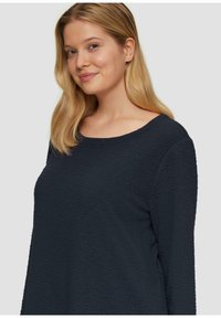 Jonge vrouw met blond haar die lichtjes glimlacht, gekleed in een donkere marineblauwe top met lange mouwen tegen een effen lichte achtergrond.