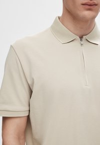 Polo beige avec un col texturé, des manches courtes et une fermeture éclair au niveau du cou. Tissu lisse avec un léger éclat.