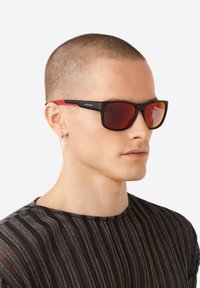 Occhiali da sole neri e rossi con lenti sfumate, forma rettangolare, finitura opaca e marchio minimale sul lato.