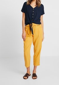 Mörkblå ribbade V-ringad blus med knyt framtill och knappar, parat med gula cropped byxor. Bruna slidessandaler med texturerad ovandel.