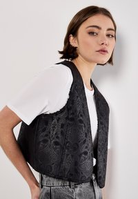 Giovane donna in una camicia bianca a maniche corte e un gilet nero a motivo paisley, in posa di lato contro un muro bianco semplice.