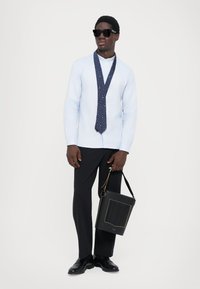 Mężczyzna w jasnoniebieskiej koszuli na guziki, z rozwiązaną granatową krawatem w wzory, czarnych spodniach, czarnych butach, okularach przeciwsłonecznych i trzymający czarną torbę.