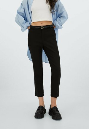Kobieta ubrana w czarne dopasowane spodnie z czarnym paskiem, biały krótki top, niebieską oversize koszulę oraz czarne masywne loafersy, stoi z rękami za plecami.