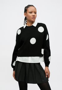 Μαύρο πουλόβερ με μεγάλα λευκά πουά, στρογγυλή λαιμόκοψη, άνετη εφαρμογή, φορεμένο πάνω από λευκό πουκάμισο με γιακά και γκρι πλισέ φούστα.