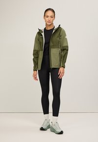 Olijfgroene waterdichte jas met een rits aan de voorkant, meerdere zakken en een capuchon. Draagt over zwarte legging en lichte groene sportschoenen.