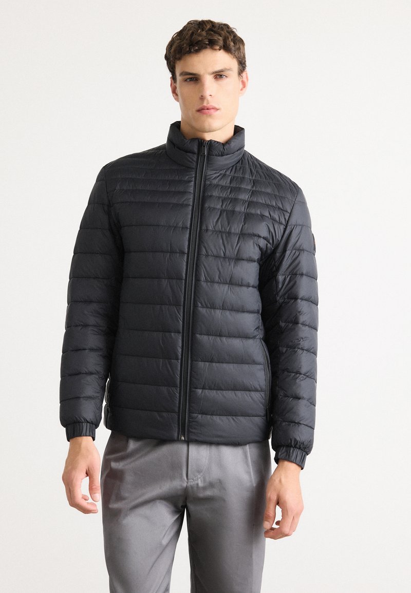 Young man wearing a black quilted zip-up jacket and light gray pants, standing against a plain white background.