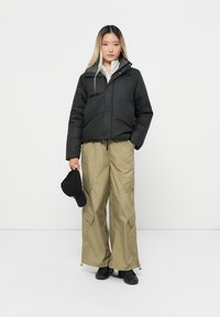 Zwarte pufferjack met een hoge kraag, gedragen over een grijze top, gecombineerd met losse kaki broeken en zwarte schoenen. Houdt een zwarte hoed vast.