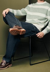 Gestreifter Pullover in Grün und Weiß, dunkle Denim-Jeans und braune Schuhe mit Gummisohlen. Person sitzt in einem schwarzen Drahtstuhl, die Beine crossed.