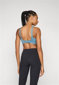 Mujer con cabello trenzado vistiendo un sujetador deportivo azul y leggings negros de cintura alta, de pie con la espalda hacia la cámara.
