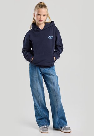 Felpa blu navy con tasca frontale e cappuccio regolabile, abbinata a jeans denim blu a gamba larga e sneakers grigie.