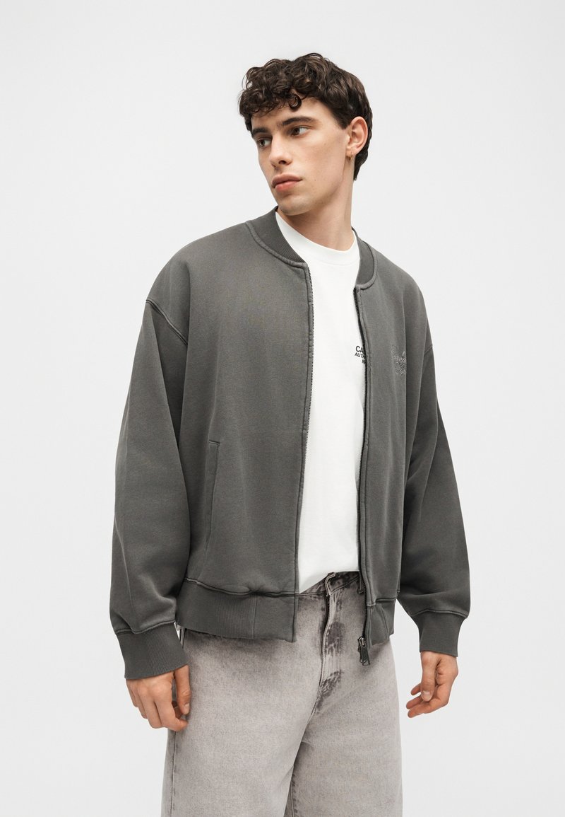 Joven con el pelo rizado y corto que lleva una chaqueta gris oscuro con cremallera sobre una camiseta blanca y vaqueros gris claro, mirando de lado.