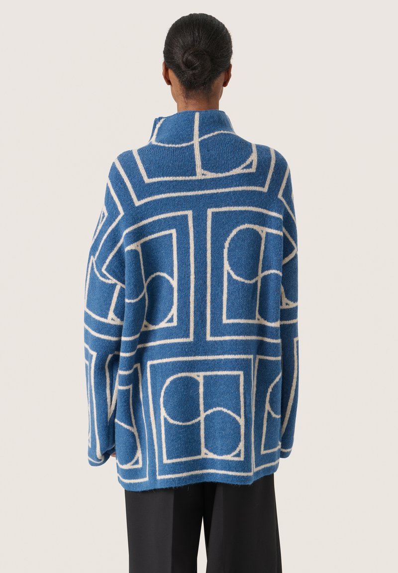 Blauer Wollpullover mit geometrischem Muster in Creme. Auffällige Merkmale sind der hohe Kragen und die oversized Passform sowie die weiche Textur.