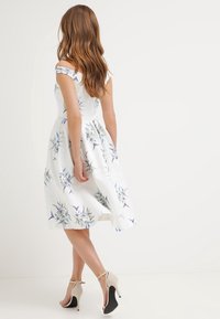 Robe blanche à motif floral, épaules dénudées, avec une jupe évasée et une surface texturée. Portée avec des chaussures à talons hauts couleur nude.
