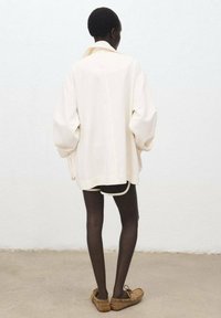 Oversized crèmekleurige jas met verlaagde schouders, achterkantnaaddetail en ribbelkraag, gecombineerd met zwarte leggings en bruine schoenen.