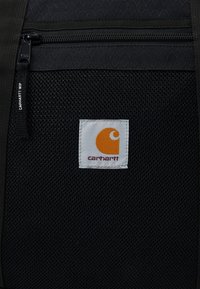 Czarna teksturowana tkanina z zamkiem błyskawicznym, z białą prostokątną metką z pomarańczowym logo i napisem "carhartt" w ciemnoczerwonym kolorze.