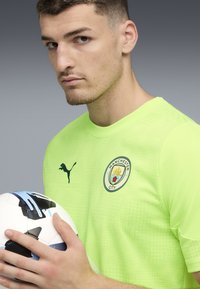 Neonvihreä urheiluasu, jossa on Puma-logo ja Manchester City -merkki. Pelaaja pitää mustavalkoista jalkapalloa, jossa on sinisiä yksityiskohtia.