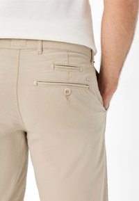 Pantaloni beige realizzati in tessuto liscio, con due tasche posteriori dotate di chiusure a bottone e una minimale etichetta sul retro.