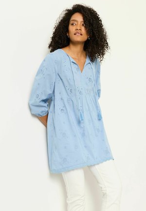 Žena s kučeravými vlasmi, oblečená v svetlomodrej vyšívanej tunike s strapcami, stojaca na jednolnom svetlom pozadí.