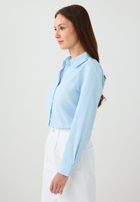 Woman with long brown hair wearing a light blue buttoned shirt and white pants, standing sideways against a plain background.