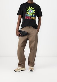 Camiseta de algodón negra con un colorido diseño gráfico de un sol, combinada con pantalones beiges holgados y zapatillas de color crema, sosteniendo una bolsa negra.