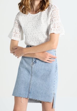 Personne portant un haut blanc en dentelle à manches courtes et une jupe en denim bleu clair avec un ourlet brut et une fente latérale, debout les bras croisés.