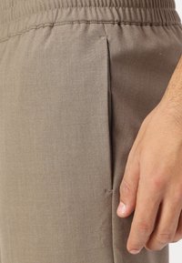 Pantaloni in tessuto beige con vita elasticizzata, dotati di una tasca laterale e una sottile trama. Inquadratura ravvicinata del pantalone e della mano.