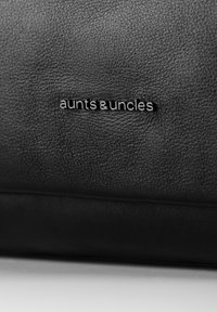 Zwarte, gestructureerde leren oppervlak met een klein metalen logo met de tekst "aunts & uncles" gecentreerd in het beeld.