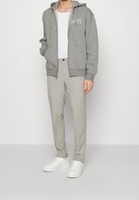 Sweat à capuche gris zippé avec une poche kangourou, associé à un pantalon beige clair et des baskets blanches. Design simple avec cordons de serrage et logo.