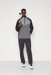 Set di abbigliamento da golf composto da un pullover grigio e nero con mezza zip e pantaloni scuri abbinati. Indossato con scarpe da golf e impugnando una mazza da golf.