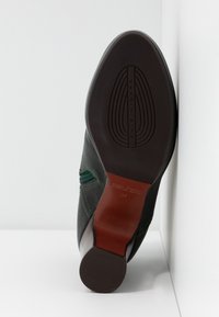 Chaussure habillée noire à semelle cuir, talon en caoutchouc, coutures visibles et logo de marque embossé sur la semelle, inclinée contre un coin blanc.