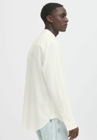 Camisa de manga larga, de color beige claro, en un corte relajado, con cuello abotonado y dobladillo curvado, que presenta una tela suave y texturizada.