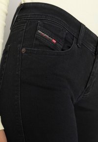 Jeans de mezclilla negra con un ajuste slim, que presentan un pequeño bolsillo para monedas con una etiqueta de logo roja y blanca, costuras visibles y una textura suave.