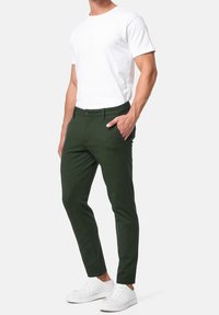 Chinos ajustés verts en tissu lisse, avec poches latérales et fermeture par bouton. Portés avec un t-shirt blanc uni et des baskets blanches.