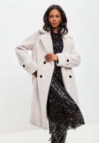 Abrigo gris claro de doble botonadura, hecho de una tela suave, con botones negros y solapas oversize, combinado con un vestido negro de lentejuelas.