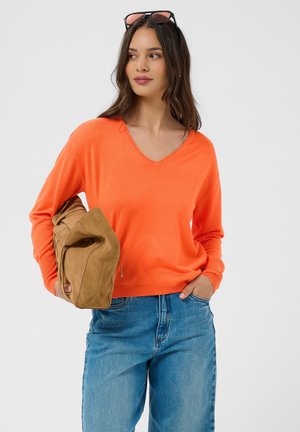 Vrouw in oranje shirt met lange mouwen en blauwe spijkerbroek houdt een bruine suède tas vast en draagt een zonnebril op haar hoofd tegen een effen achtergrond.