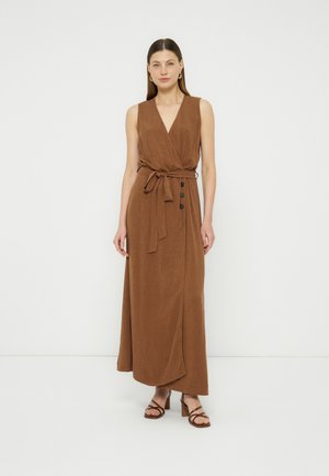 Frau trägt ärmelloses braunes Wickelkleid mit Taillengürtel und Knöpfen, kombiniert mit braunen, Riemchen-High-Heel-Sandalen, vor weißem Hintergrund stehend.