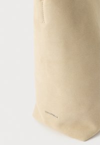 Sac à main en suède beige avec détails de couture subtils et petit logo argenté "COCCINELLE" près du bas sur fond blanc.