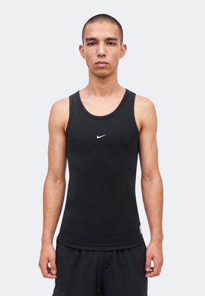 Jeune homme aux cheveux courts portant un débardeur noir ajusté Nike et un short noir, debout devant un fond clair uni.