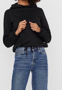 Schwarzer Kapuzenpullover mit elastischen Bündchen, kombiniert mit blauen, hoch taillierten Jeans mit Frontbindedetail und klassischem Fünf-Taschen-Design.